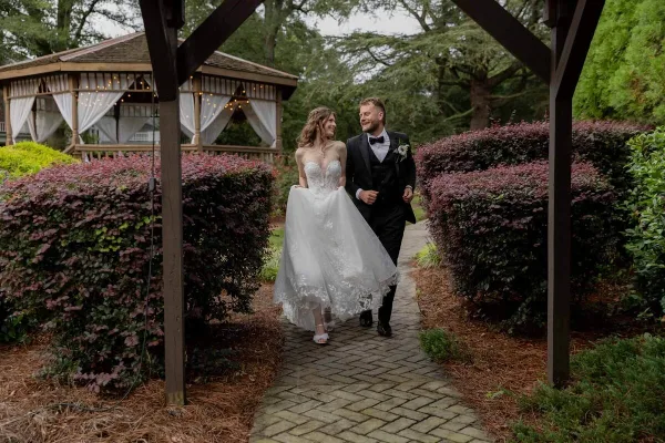 A bride in a white lace gown and a groom in a black tuxedo walking hand-in-hand along a brick pathway in a garden.