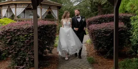A bride in a white lace gown and a groom in a black tuxedo walking hand-in-hand along a brick pathway in a garden.
