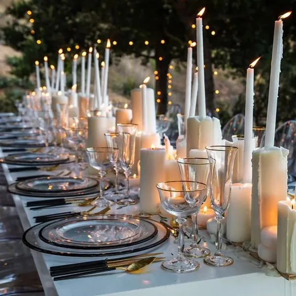 An elegant outdoor tablescape featuring a long row of white taper candles, glassware, and silver-rimmed plates.