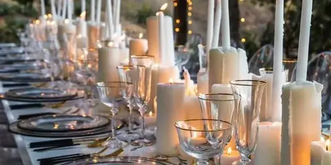 An elegant outdoor tablescape featuring a long row of white taper candles, glassware, and silver-rimmed plates.