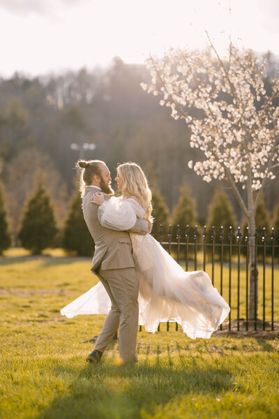 A bride and groom embracing in a grassy field with a blooming tree in the background.
