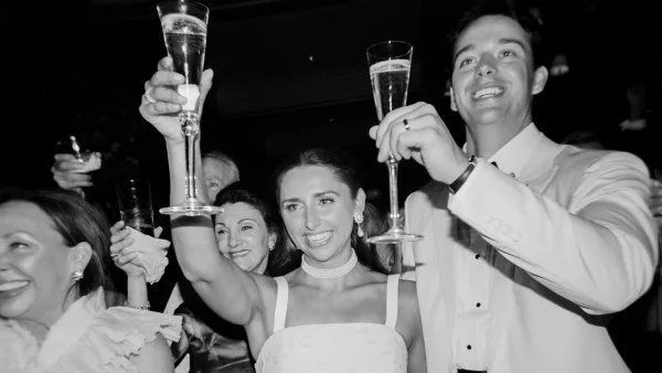 Black-and-white photo of a bride and groom raising champagne flutes in a toast at a wedding reception.