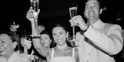Black-and-white photo of a bride and groom raising champagne flutes in a toast at a wedding reception.