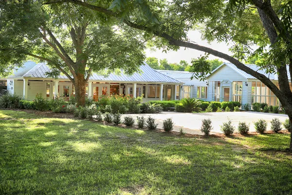 Exterior view of a white wooden banquet hall with a metal roof, surrounded by green lawn, trees, and landscaping.