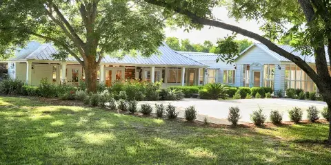 Exterior view of a white wooden banquet hall with a metal roof, surrounded by green lawn, trees, and landscaping.