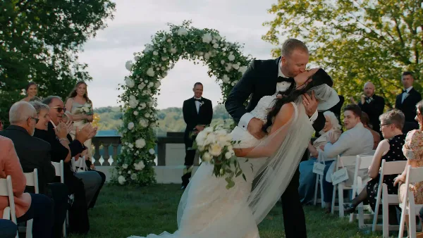 A groom in a tuxedo lifts his bride in a kiss during an outdoor wedding ceremony under a floral arch.