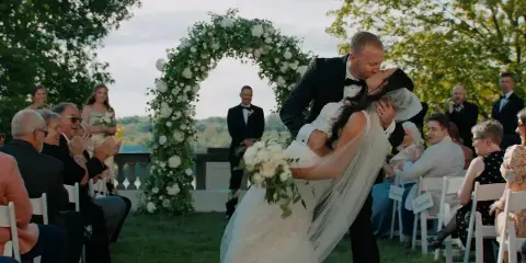 A groom in a tuxedo lifts his bride in a kiss during an outdoor wedding ceremony under a floral arch.