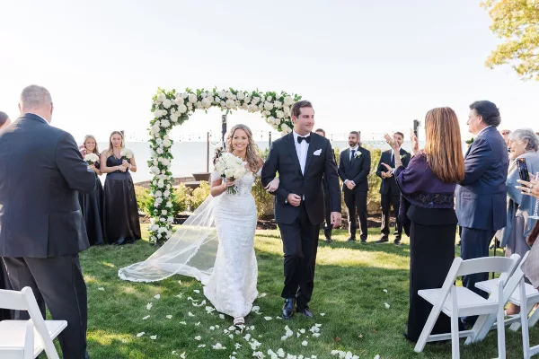 A bride and groom walking down the aisle at an outdoor wedding ceremony with guests and a floral arch.