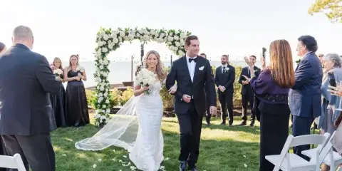 A bride and groom walking down the aisle at an outdoor wedding ceremony with guests and a floral arch.