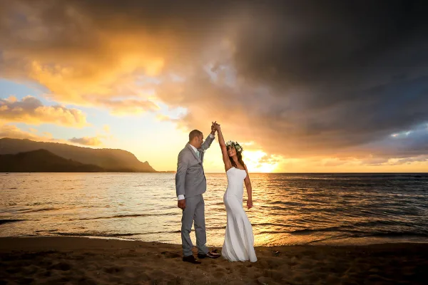 A bride and groom dancing on a sandy beach at sunset with the ocean and mountains in the background.