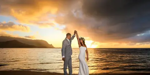 A bride and groom dancing on a sandy beach at sunset with the ocean and mountains in the background.