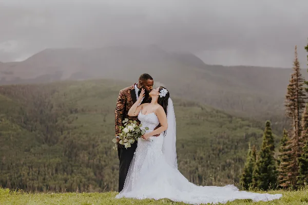A bride and groom sharing a kiss on a grassy hill with a mountain range in the background.