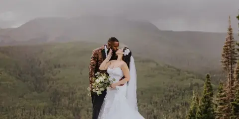 A bride and groom sharing a kiss on a grassy hill with a mountain range in the background.