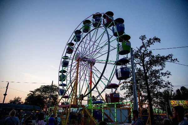 A large illuminated Ferris wheel at a fairground event with visitors and trees at dusk.