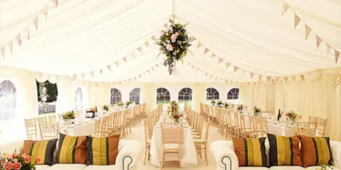Interior of a white wedding tent featuring long reception tables, chairs, and floral decorations.