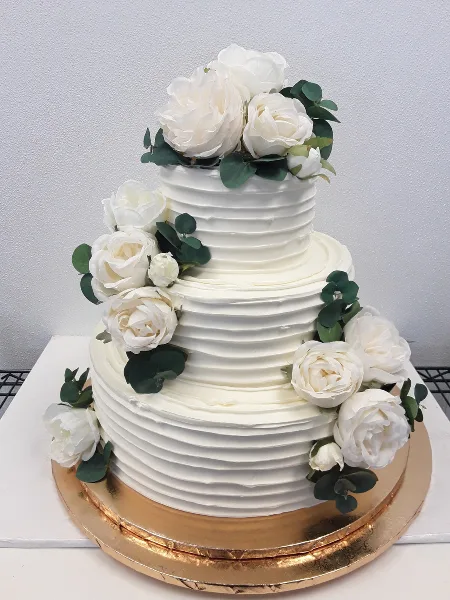 A three-tier white wedding cake with textured buttercream frosting, decorated with white roses and eucalyptus leaves, sitting on a gold cake board.