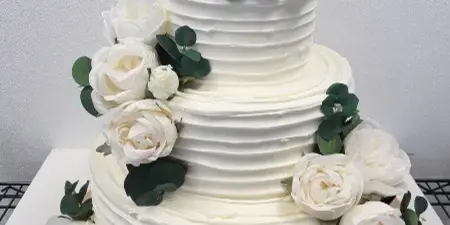 A three-tier white wedding cake with textured buttercream frosting, decorated with white roses and eucalyptus leaves, sitting on a gold cake board.