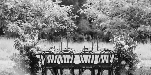 A black and white photograph of a dining table surrounded by bentwood chairs, set up outdoors with floral arrangements.