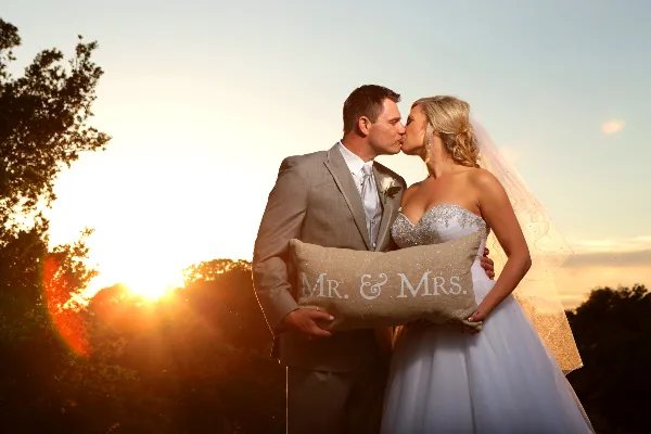 A bride and groom kissing while holding a 'Mr. & Mrs.' pillow against a sunset background.