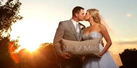 A bride and groom kissing while holding a 'Mr. & Mrs.' pillow against a sunset background.