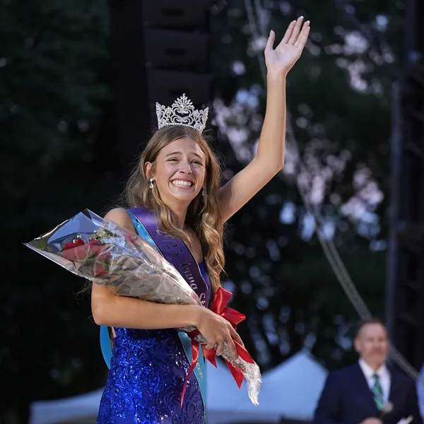 A young woman wearing a crown and sash waves to a crowd while holding a bouquet of red roses.