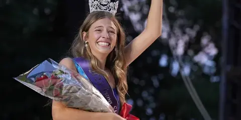 A young woman wearing a crown and sash waves to a crowd while holding a bouquet of red roses.