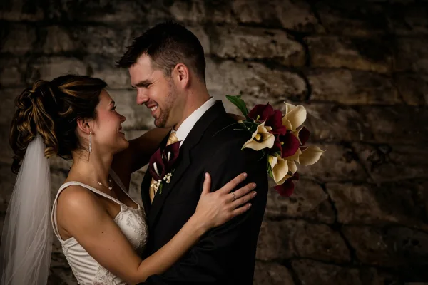 A bride and groom embracing and smiling in front of a stone wall, with the bride holding a bouquet of calla lilies.