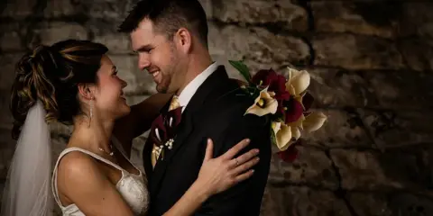 A bride and groom embracing and smiling in front of a stone wall, with the bride holding a bouquet of calla lilies.