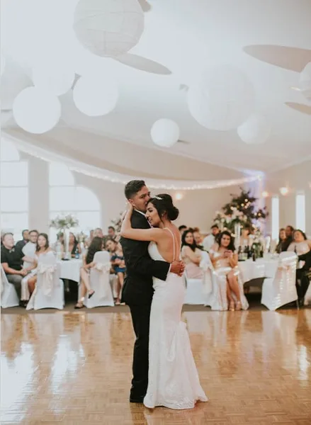 A bride and groom dancing during a wedding reception in a large ballroom with guests seated at tables.
