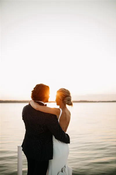 A bride and groom embracing at sunset by a body of water.