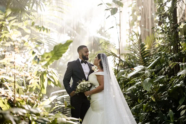 A bride and groom standing together in a lush, sunlit garden setting.