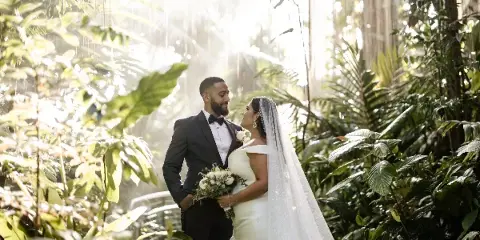 A bride and groom standing together in a lush, sunlit garden setting.