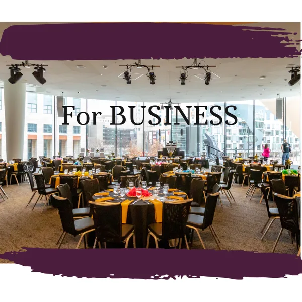 A large event venue with round tables set for a formal dinner, featuring black tablecloths, yellow napkins, and stage lighting.