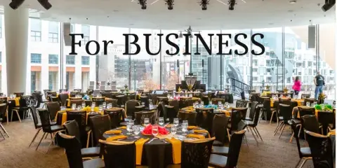 A large event venue with round tables set for a formal dinner, featuring black tablecloths, yellow napkins, and stage lighting.