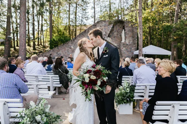 A bride and groom kissing during an outdoor wedding ceremony in a wooded area with guests seated on white benches.
