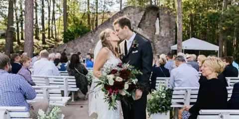 A bride and groom kissing during an outdoor wedding ceremony in a wooded area with guests seated on white benches.