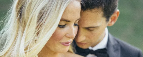A close-up photograph of a bride and groom in wedding attire, with the groom resting his head near the bride's.