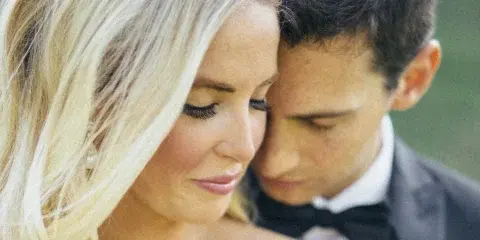 A close-up photograph of a bride and groom in wedding attire, with the groom resting his head near the bride's.