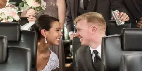 Bride and groom smiling at each other with bridal party holding bouquets inside a limousine.
