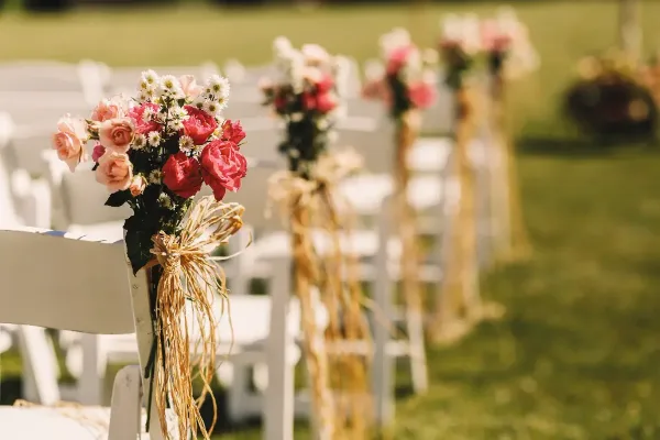Outdoor wedding ceremony aisle with white chairs decorated with pink and red roses and burlap ties.