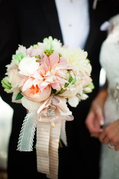 A close-up of a bridal bouquet with pink and white flowers and ribbons held by a couple.