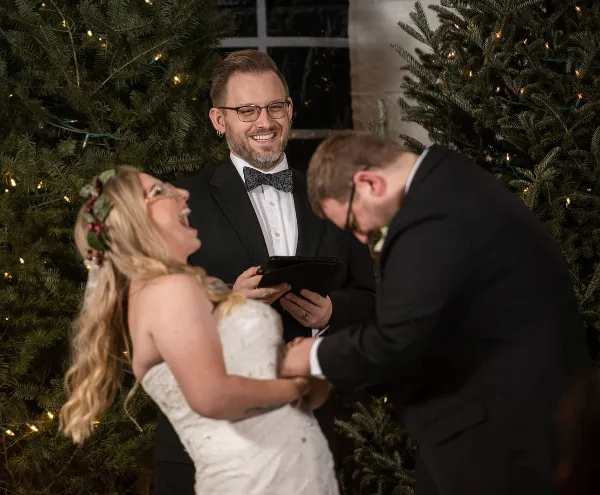 A smiling officiant in a tuxedo holding a tablet while officiating a wedding ceremony for a bride and groom.
