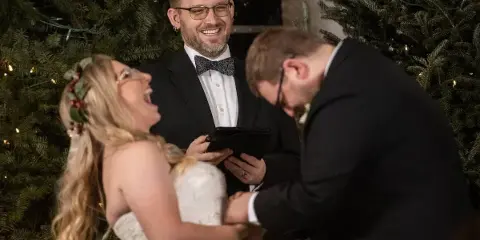 A smiling officiant in a tuxedo holding a tablet while officiating a wedding ceremony for a bride and groom.