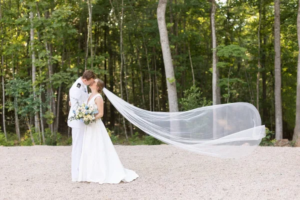 A bride and groom sharing a kiss in a wooded area, with the bride's long veil blowing in the wind.