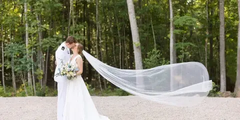A bride and groom sharing a kiss in a wooded area, with the bride's long veil blowing in the wind.