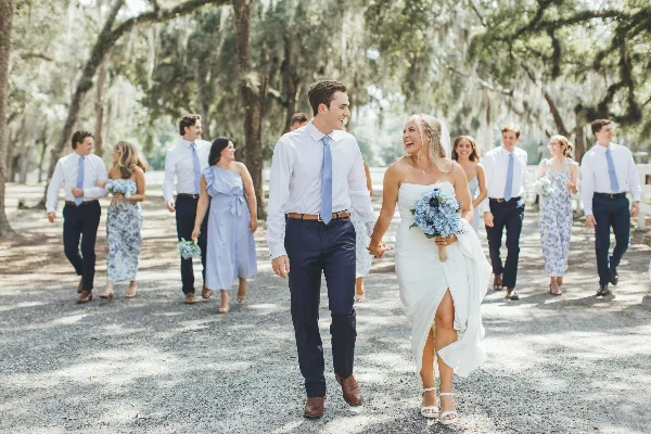 A bride and groom walking hand-in-hand with their wedding party in a wooded outdoor setting.
