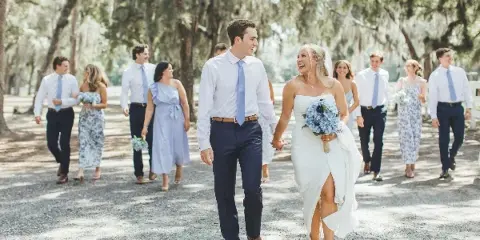 A bride and groom walking hand-in-hand with their wedding party in a wooded outdoor setting.