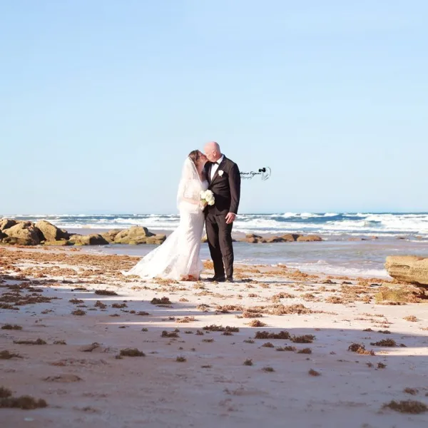A bride and groom standing on a sandy beach with the ocean in the background.