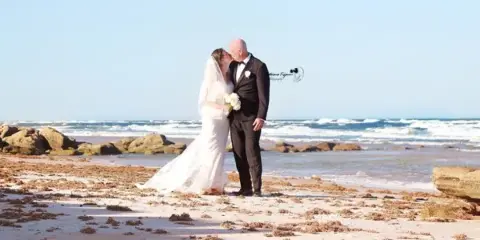 A bride and groom standing on a sandy beach with the ocean in the background.