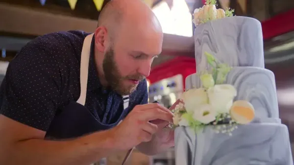 A baker in an apron carefully placing white flowers onto a grey marbled wedding cake.